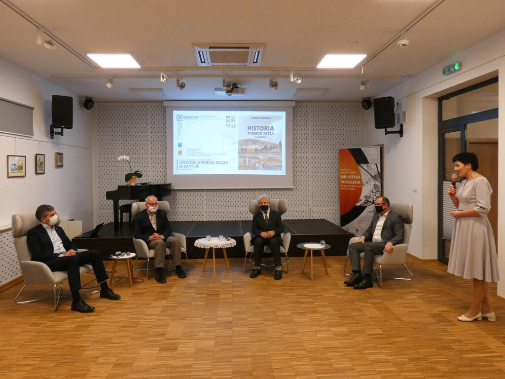 Na zdjęciu pięć osób na sali: cztery osoby siedzą jedna przemawia